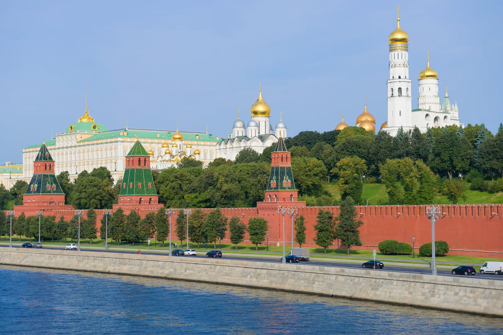 The Moscow Kremlin from across the Moskva River on a sunny day: the red brick wall and towers, the Grand Kremlin Palace and the gold domes of the cathedrals behind.